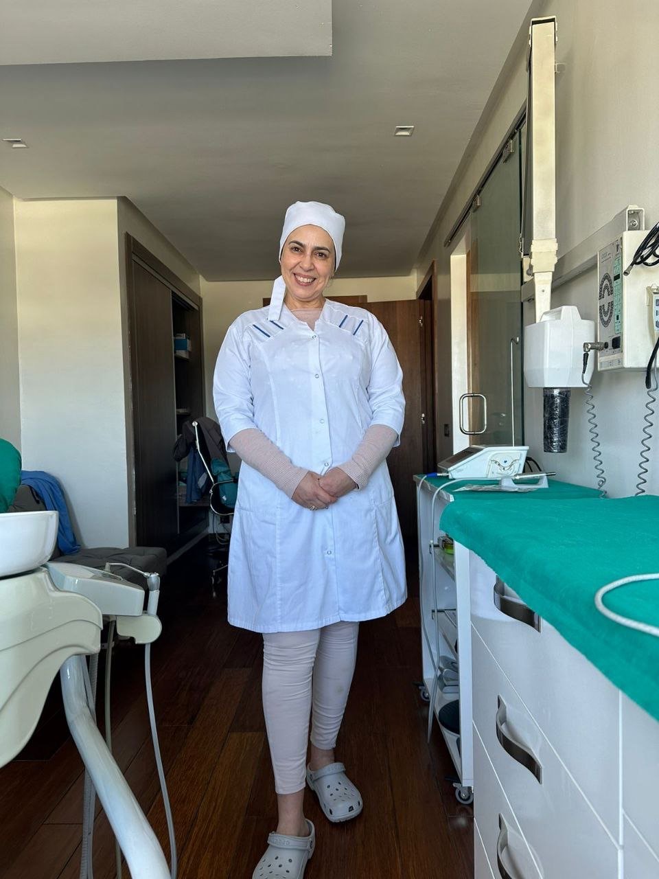Praticienne souriante en blouse blanche, debout dans une salle de soins lumineuse du centre, entre le fauteuil dentaire et le plan de travail